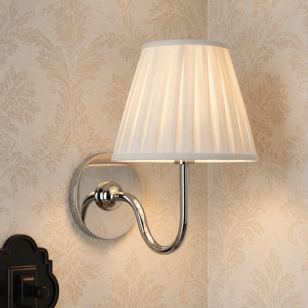 Close view of a Tansy single-light sconce in Chrome: round mounting plate, curved arm and pleated linen shade against patterned wallpaper